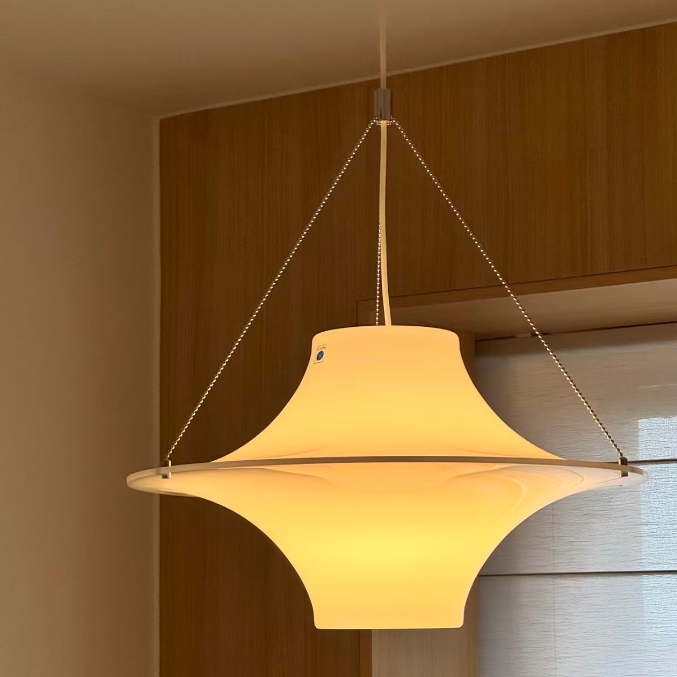 A modern mid-century style pendant light with a warm glowing cream-colored shade, suspended by thin metal chains in an indoor setting with wooden paneling.