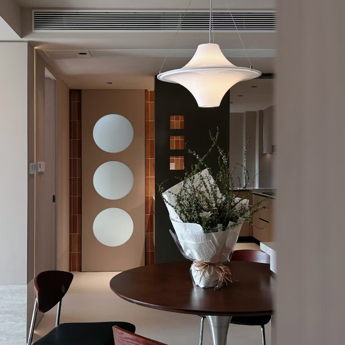A modern dining area featuring a round dark wood table with a silver pedestal base, surrounded by contemporary chairs with black seats and reddish-brown wooden backs. A white dome pendant light hangs overhead, and a wrapped floral arrangement sits on the table.