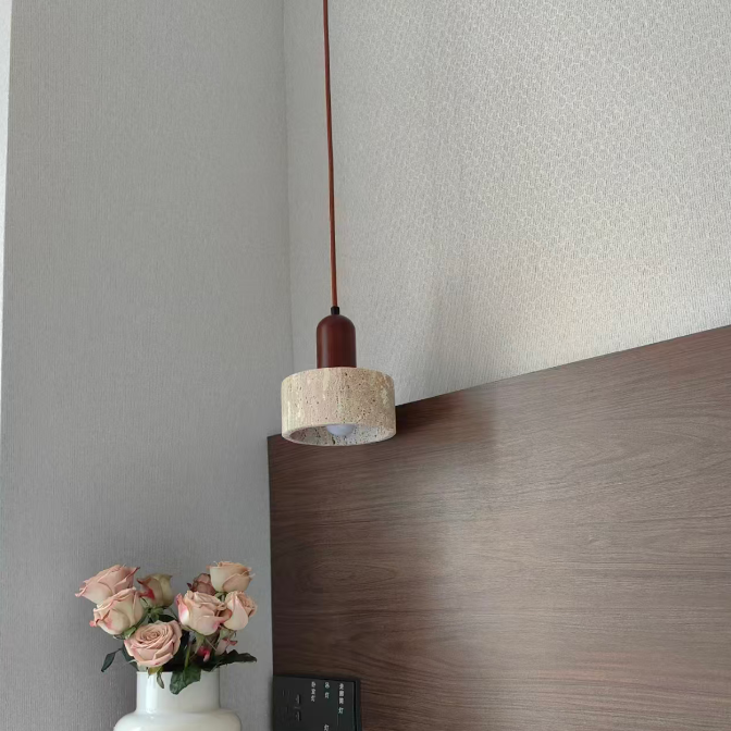 A modern pendant light with a textured beige drum shade and a dark reddish-brown wooden cap, hanging in a bedroom setting above a dark wood headboard.