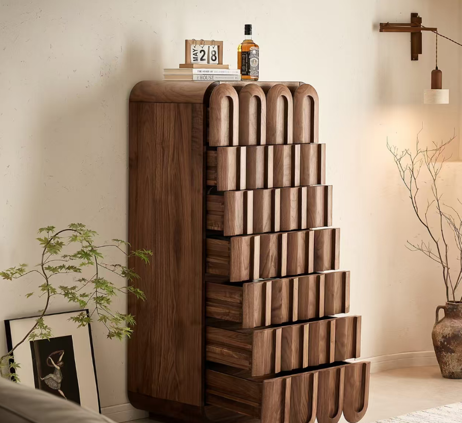 A tall walnut wood chest of drawers with a distinctive arched drawer front, styled in a warm, modern room setting next to a plant and lamp.