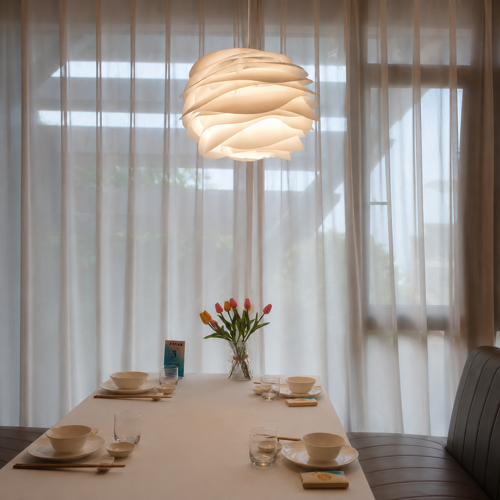 Dining table set with a vase of flowers under a modern pendant light, with sheer curtains in the background.