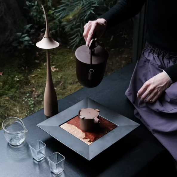Person pouring tea from a black teapot into a glass on a dark surface with a blurred natural background