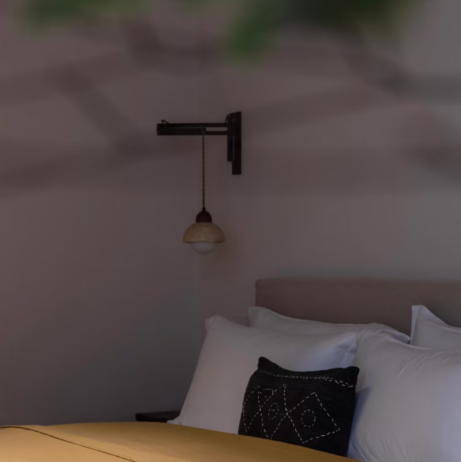 A modern black wall-mounted sconce with a conical shade hanging above a bed with white pillows and a mustard yellow duvet in a dimly lit bedroom.
