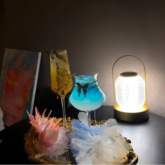 Decorative setup with a lit lamp, cocktail glass, and flowers on a dark surface.