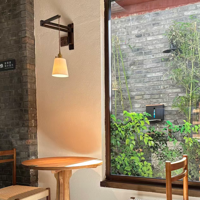 A cozy cafe corner with a round wooden table, two wooden chairs with woven seats, a wall-mounted lamp, and a large window looking out onto green plants and a gray brick wall.