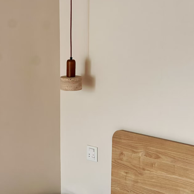 A minimalist bedroom corner featuring a modern pendant light with a dark brown cord and cylindrical shade, a light wood headboard, white bedding, and a small woven side table against a warm beige wall.